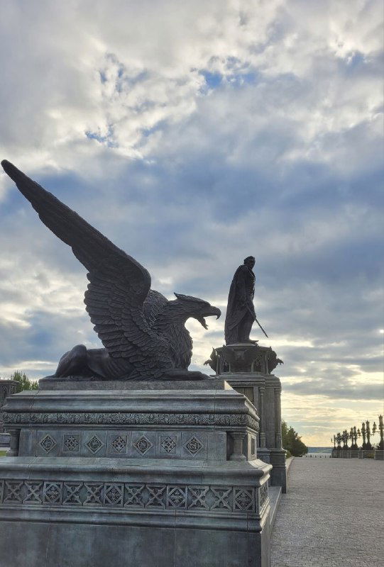Силуэты скульптуры крылатого существа и фигуры на пьедестале на фоне рассеянного неба и набережной Волги