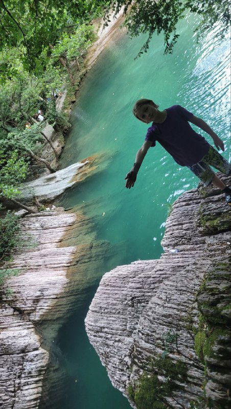 Человек стоит на выступающей белой скале у берега прозрачной воды, на заднем плане густая зелёная растительность и тихая заводь с отражениями.