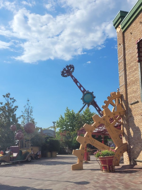 Декоративные большие шестерёнки и элементы паровой машины у стены здания; вокруг — горшки с растениями и пешеходная зона в парке развлечений.