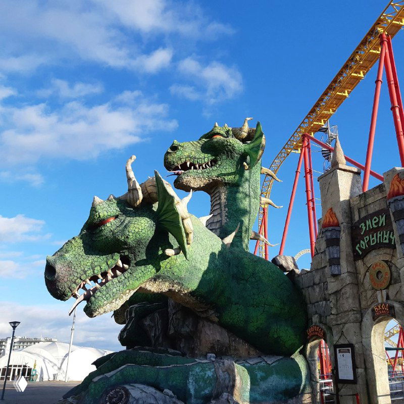 Скульптура зелёного трёхглавого Горыныча на фоне аттракционов; крупный план тематического персонажа возле входа в развлекательную зону.
