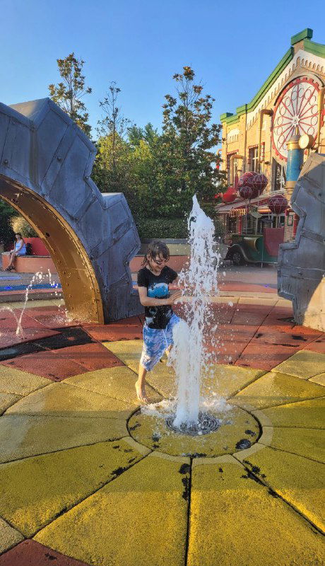 Ребёнок стоит в центре струи фонтана на ярком круглом покрытии детской площадки рядом с большой декоративной шестерёнкой; летний день и зелень позади.