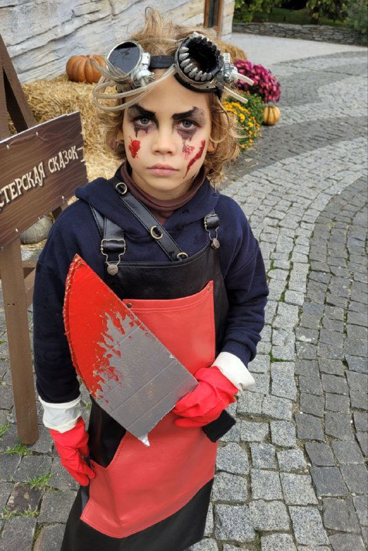 Крупный портрет ребёнка в хэллоуинском образе: мэйк с тёмными глазами, гогглы на лбу, красный фартук и большой нож-бутафория на фоне мощёной площади.