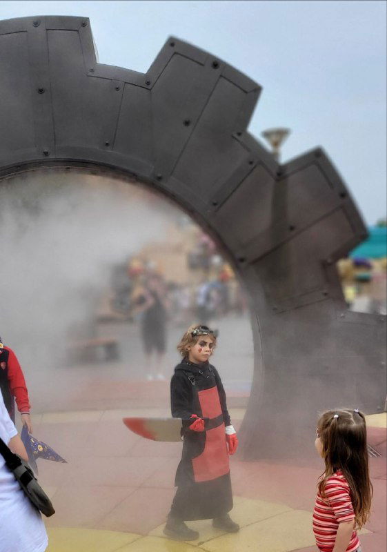 Ребёнок в костюме идёт через туманную зону у той же большой шестерёнки; вокруг размытые прохожие и атмосферный фон парка развлечений.