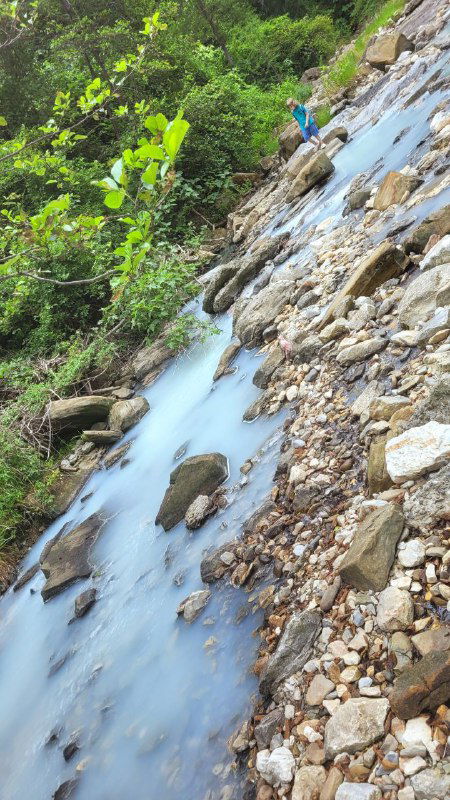 Каменистый склон и поток бирюзовой воды, вода стекает между валунами, берег зарос эвкалиптами и кустарником вблизи минерального источника