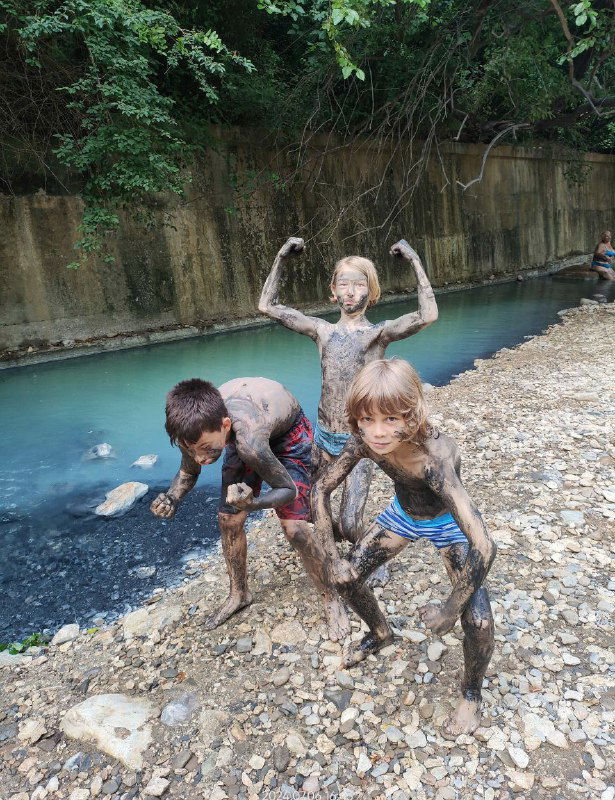Дети, покрытые лечебной грязью, стоят на берегу бирюзовой реки Мацеста — игровой и лечебный момент у минерального источника