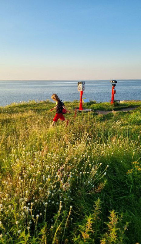 Пляжный прибрежный склон с травой и красными сигнальными опорами, ребёнок бежит по траве на фоне моря и горизонтального закатного света.