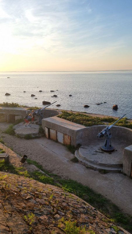 Панорамный вид на оборонительные сооружения у моря: небольшой причал, камни, артиллерийские позиции и спокойная морская гладь в золотой час.