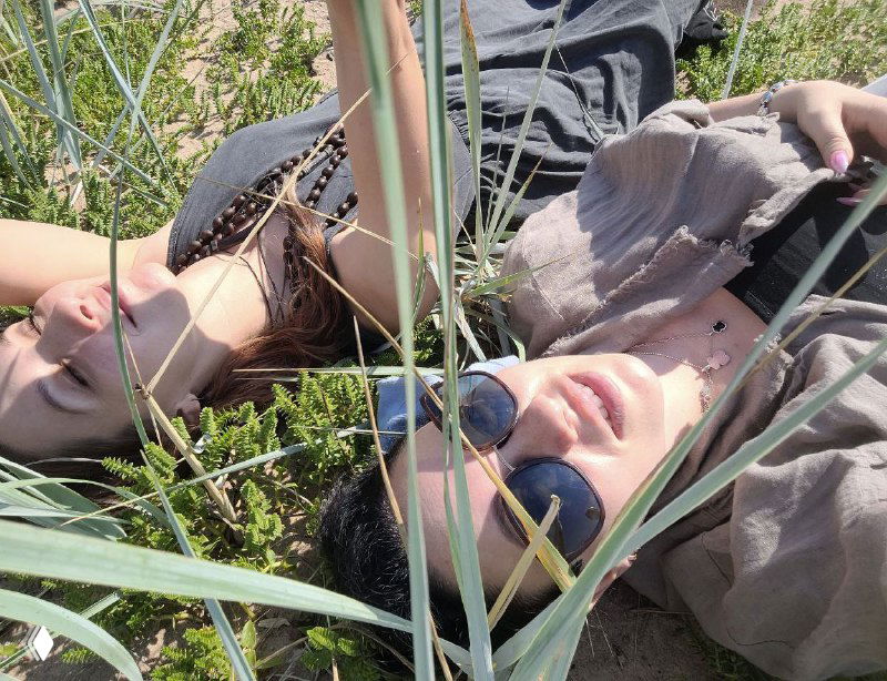 Двое взрослых лежат в траве, снимают момент отдыха на природе: солнце, зелёные стебли и ощущение ленивого летнего вечера у моря.