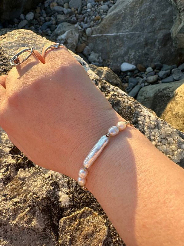 Запястье вблизи с удлинённой жемчужиной и круглыми бусинами на фоне прибрежных скал — текстура кожи и металла украшений видна чётко.