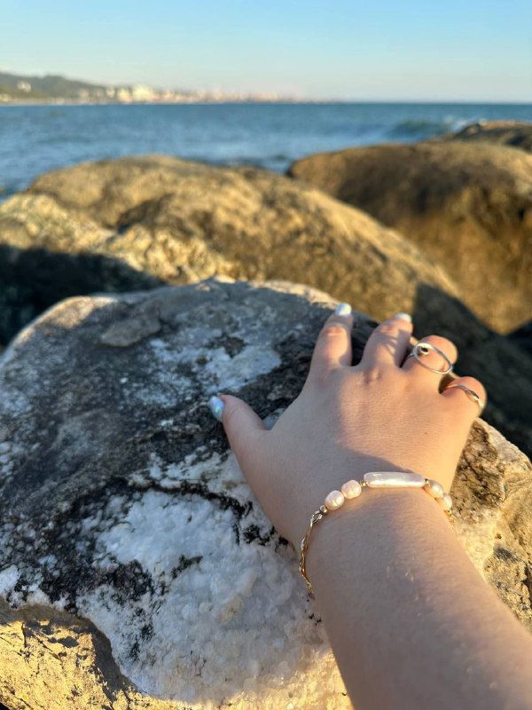 Рука с кольцами и жемчужным браслетом, поставленная на скалу у моря, кадр подчёркивает игру текстур: камень, вода и блеск украшений при закатном свете.
