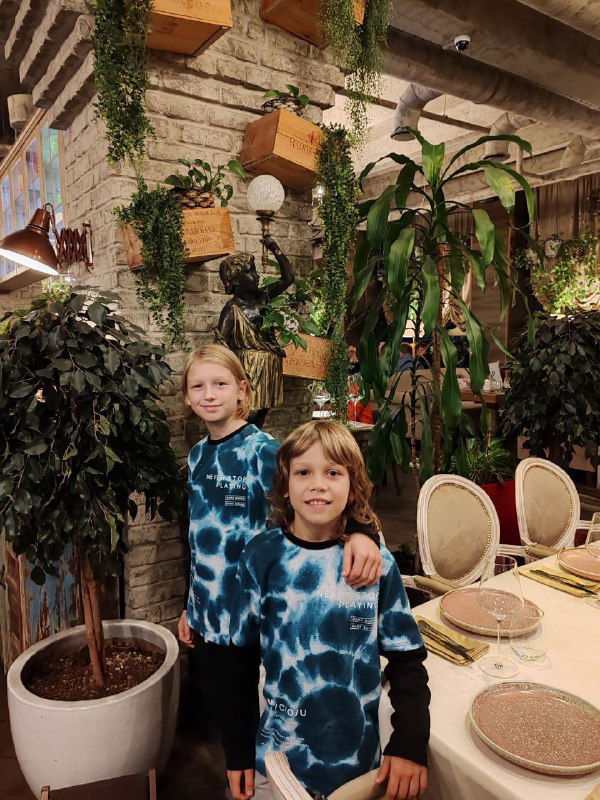 Двое детей в одинаковых тай-дай футболках в интерьере ресторана: столы, зелень и уютная обстановка заведения в Мурманске.