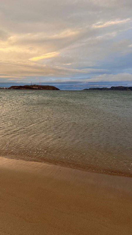 Пустынный песчаный пляж Териберки на закате, гладкая береговая линия и спокойное море под облачным небом, тёплые оттенки света.