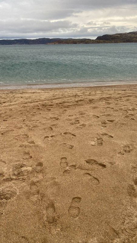 Следы ног на влажном песке пляжа Териберки, вид вверх по берегу к морю и удалённым скалам, лёгкая рябь воды.