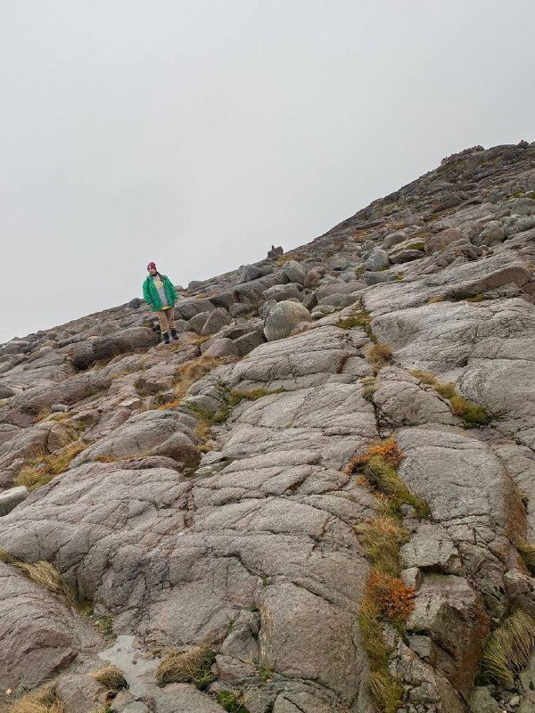 Человек в зелёной куртке поднимается по пологой скале в тундре — масштаб и трудности прогулки по каменистому берегу на фоне серого неба.