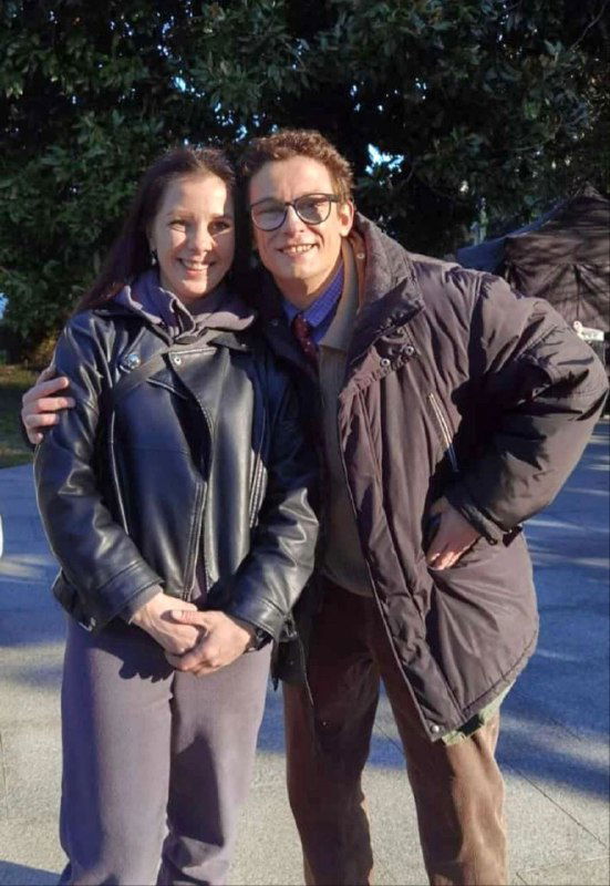 Два человека позируют вместе на улице у зелёной растительности; фото выглядит как кадр с площадки или закулисное фото съёмок.