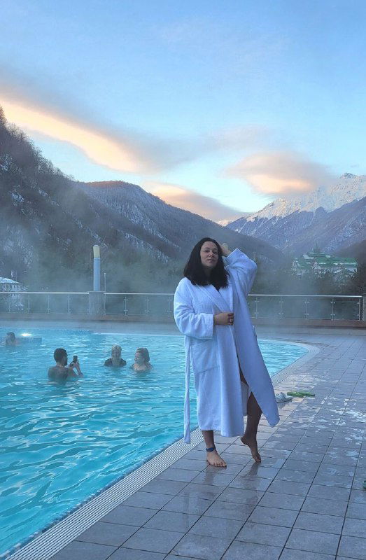 Полный рост автора в белом халате стоит у края открытого бассейна; в воде видны купающиеся люди, вокруг горы и лёгкий пар от тёплой воды, сумеречный свет.