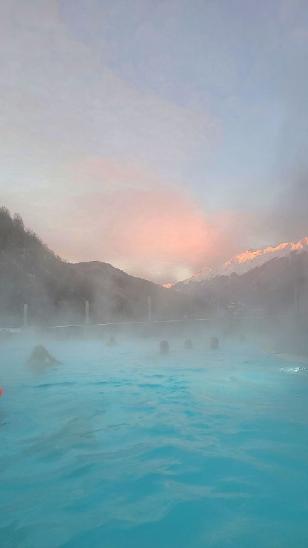 Пейзаж с паром над тёплой голубой водой открытого бассейна, вдалеке силуэты гор и розоватое небо на закате; атмосфера горного отдыха и релакса.