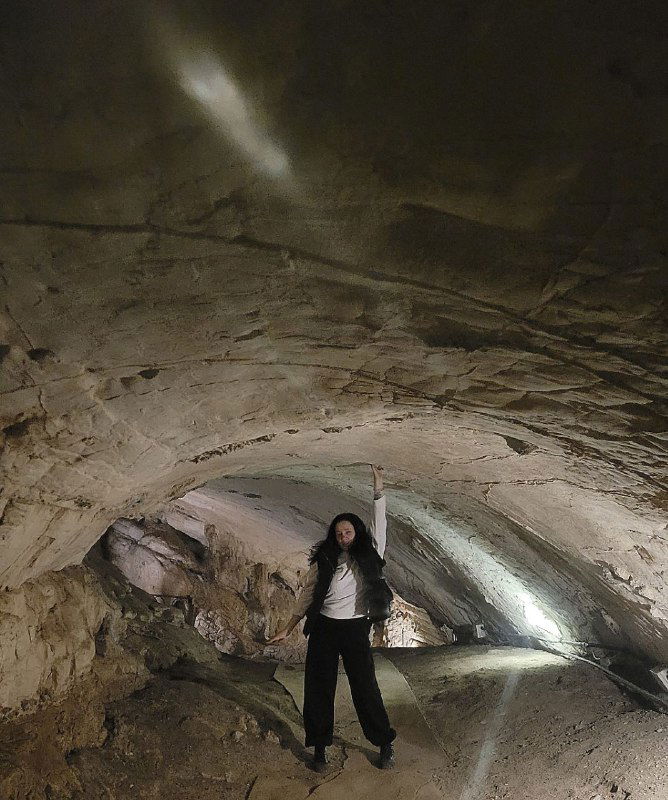 Просторная внутренняя камера пещеры с каменными сводами и мягким светом, на переднем плане силуэт человека рядом со скалой.