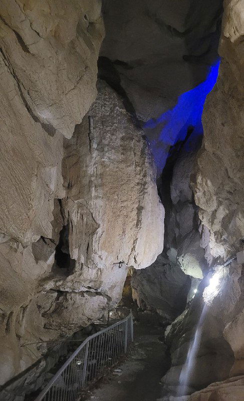 Узкий проход пещеры с каменными выступами и подсветкой, образуются вертикальные трещины и текстуры на стенах.