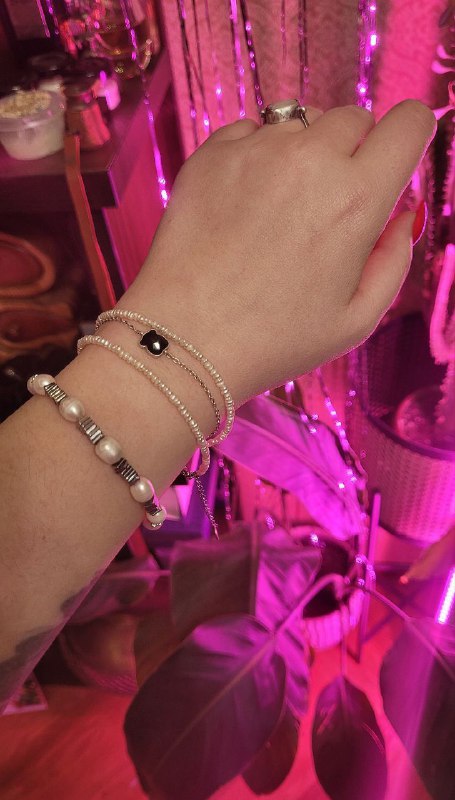 Рука с несколькими тонкими браслетами на фоне розовой подсветки и комнатных растений, видны жемчужные и цепочные модели.