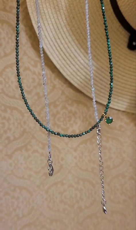 Два нежных чокера из мелких зелёных бусин и тонкой серебряной цепочки, висящие на бежевом фоне, видна подвеска и зеленый малахитовый элемент.