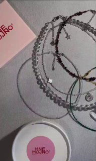 Несколько браслетов и чокеров на нейтральном фоне рядом с круглой коробочкой MNE MOJNO с розовым логотипом — видна подарочная упаковка.