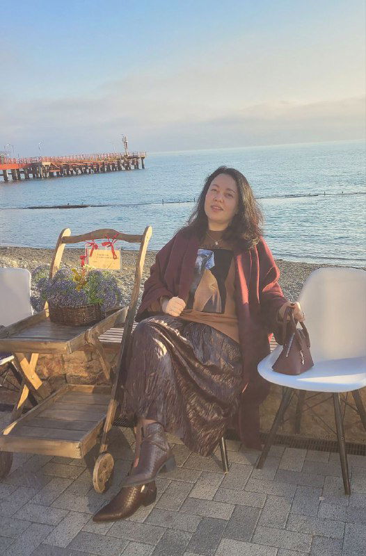 Женщина в стильной одежде сидит на набережной у моря в кафе, рядом сумка и стулья — фото личного контекста автора поста.