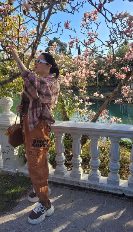 Женщина тянется к ветке с цветами тюльпановой магнолии, позируя у белой балюстрады и отражения пруда на заднем плане в солнечный день.