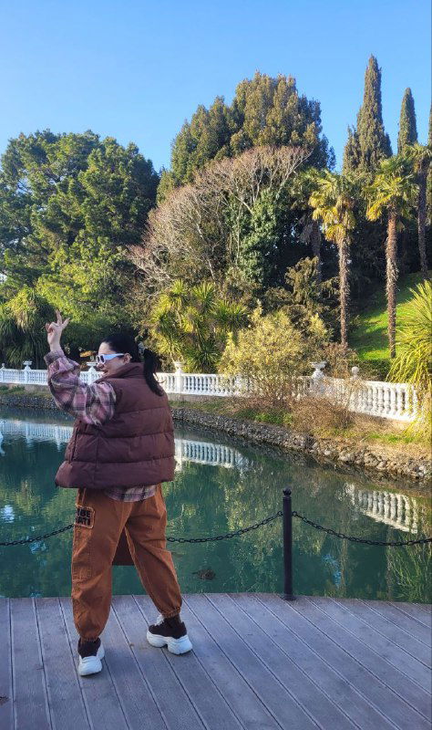 Женщина делает снимок или смотрит в камеру на деревянной платформе у пруда; вокруг видны высокие деревья и зелёные насаждения.