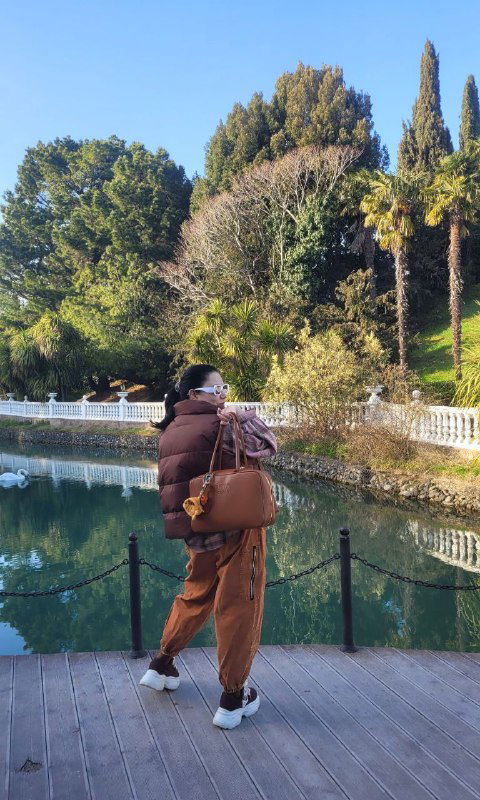 Женщина в тёплой одежде идёт по дорожке рядом с прудом; на заднем плане видны зелёные холмы и стройные деревья, создающие парковую сцену.