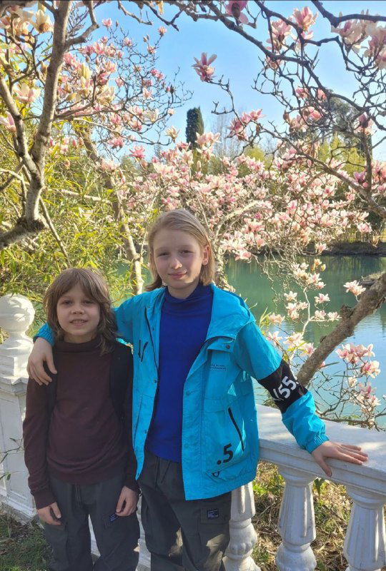 Двое детей стоят у белой балюстрады перед прудом с цветущей магнолией; детские портреты на фоне весенней зелени и отражения воды.
