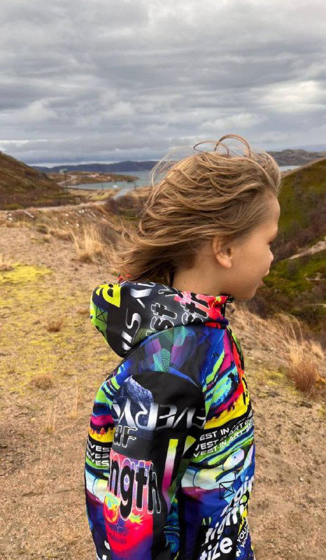 Мальчик идёт по холмистой местности в яркой разноцветной куртке; вид сзади и сбоку, природный пейзаж и влажная погода, походный кадр.