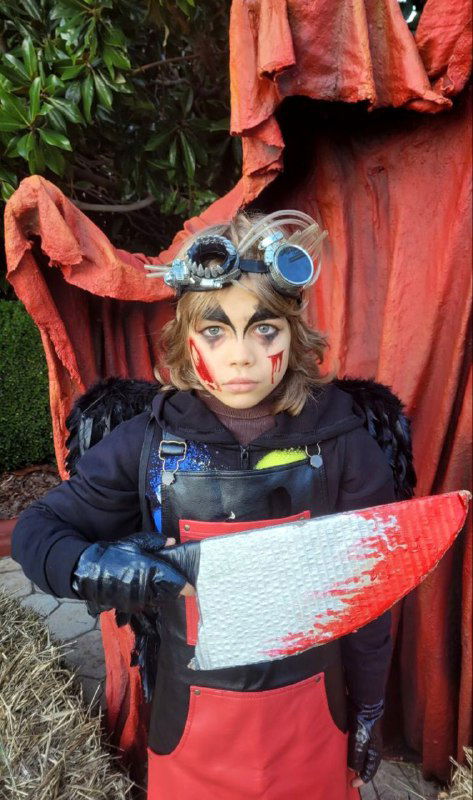 Ребёнок в карнавальном или косплейном образе с декоративным ножом; грим и костюм в театральном стиле на фоне декораций, праздничный кадр.