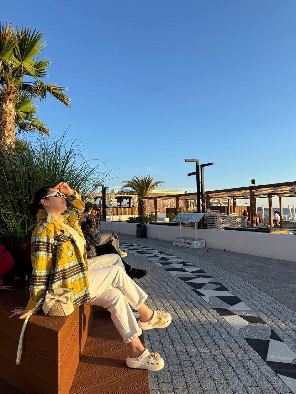 Автор отдыхает на набережной в лёгкой куртке и солнечных очках; видны лавочки, пальмы и декоративная плитка вдоль побережья курортного города.