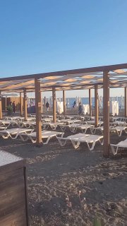 Ряд пустых шезлонгов и деревянные навесы на песчаном пляже; на заднем плане виднеется спокойное море и береговая линия курорта.