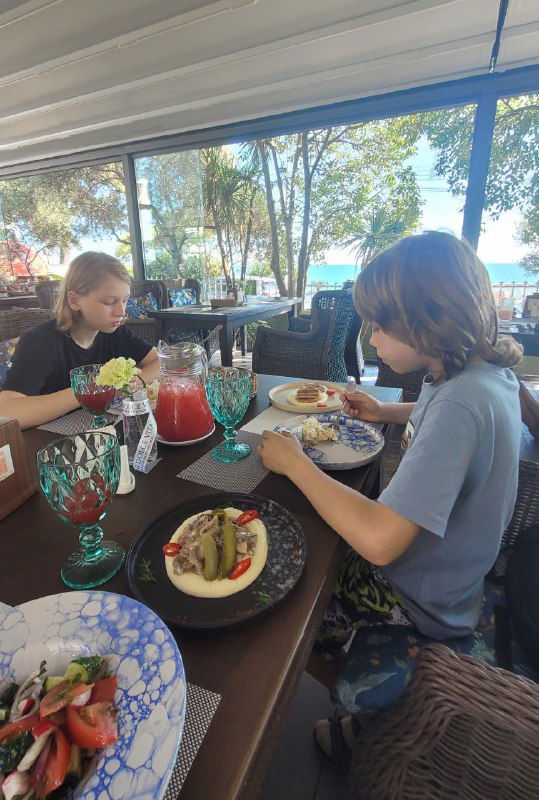 Гости ресторана за столом: блюда на тарелках, яркие бокалы и дети за обедом с видом на море и ресторанную террасу.