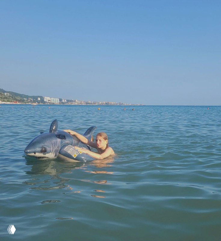 Ребёнок держится за надувную акулу в воде у берега; спокойное море, виден прибрежный пляж и гористый силуэт побережья на фоне.