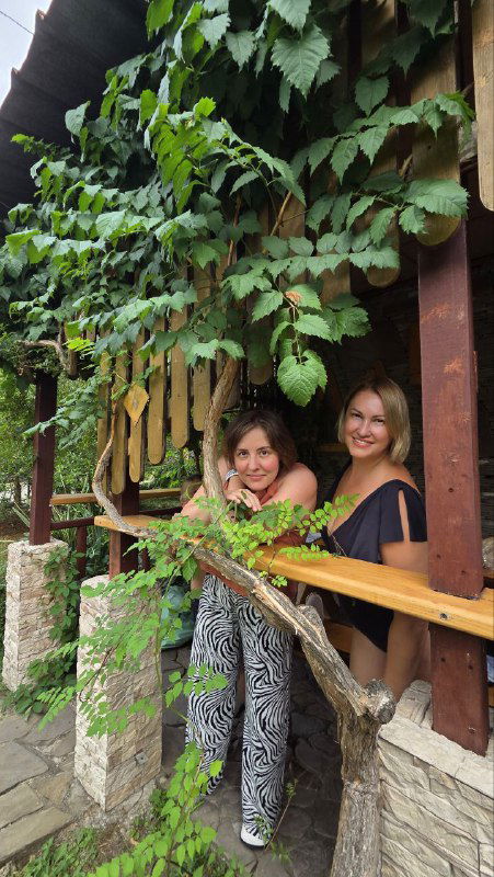 Две женщины улыбаются на балконе домика базы отдыха, вокруг вьющиеся растения и деревянные перила создают уютную сцену.