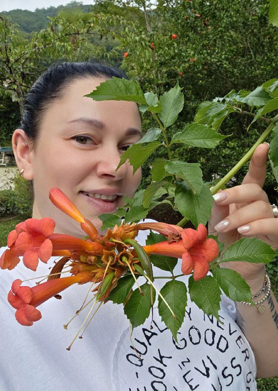 Крупный план яркого цветка в руке автора на фоне зелёной растительности базы отдыха, сочные оранжево-красные оттенки бутонов.