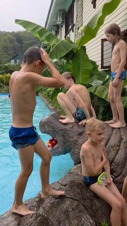 Дети на камне у бассейна базы отдыха готовятся прыгать в воду: плавки, игрушки и оживлённая летняя сцена на фоне зелени.