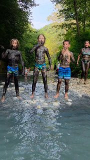 Группа детей и подростков, покрытых тёмной лечебной грязью, стоит в мелководной части реки на фоне зелени; видно одежду и счастливые лица.