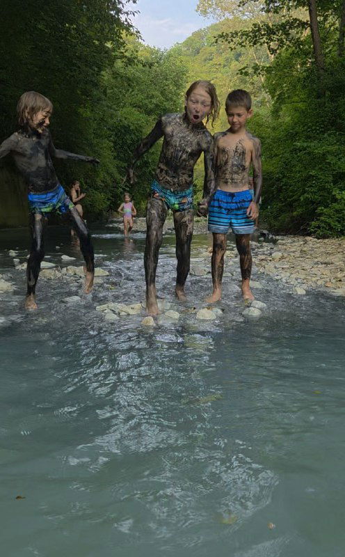 Три ребёнка, покрытые тёмной грязью, стоят в мелкой мутной воде Мацесты на фоне зелёного леса — сцена локальной игры и лечебной практики.