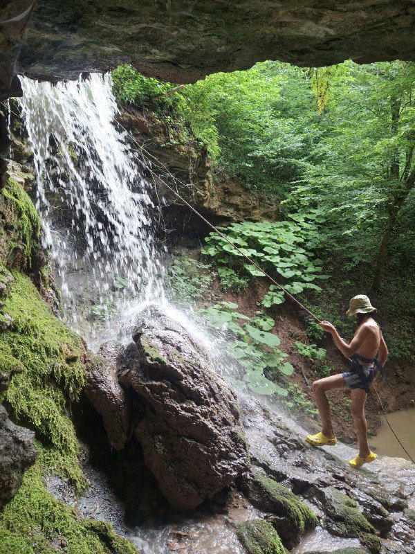 Человек у небольшого водопада в лесной горной долине: струящийся поток, влажная зелень и камни — место для природных игр и экскурсий с детьми.