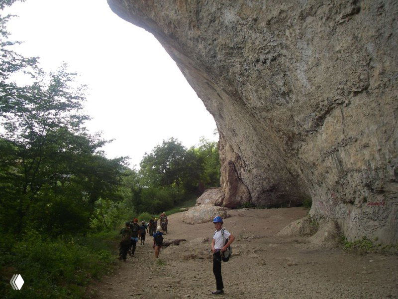 Группа людей у подножия крупного карстового свода в горах: туристы на тропе, нависающий каменный свод и лесистые склоны — подходящее место для походов и экскурсий.