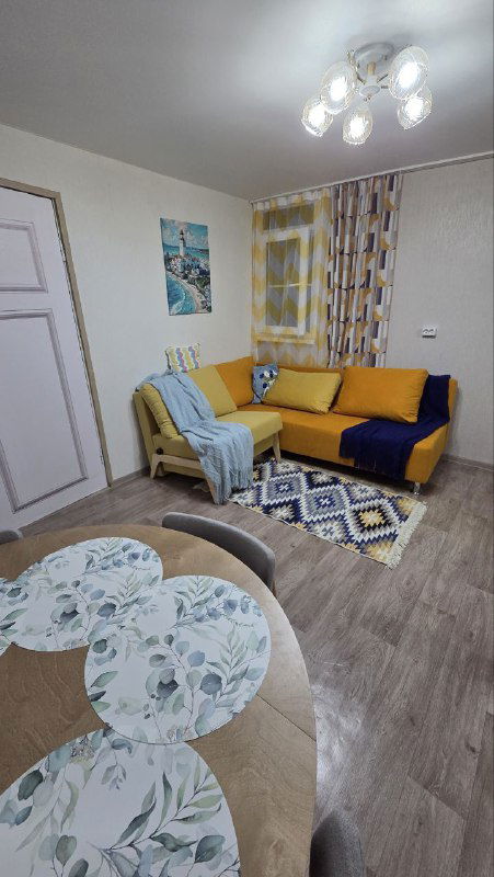 Другой ракурс той же комнаты: видна обеденная зона, жёлто-оранжевый угловой диван, занавески в клетку и потолочная люстра, деревянный пол.