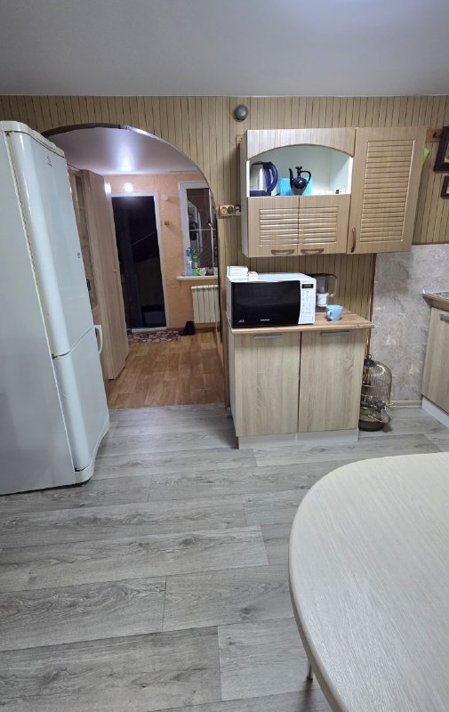 Вид на проход из кухни в прихожую: холодильник, открытая арка и элементы хранения, светлый ламинат связывает зоны в единое пространство.