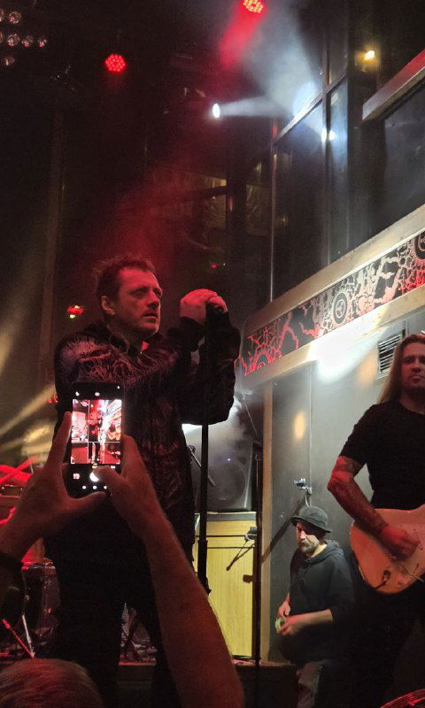 Сцена с фронтменом и сценическим монитором, в кадре также видны руки зрителей с телефонами, фиксирующими шоу с высоты зала.