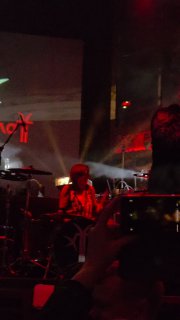 Барабанщик и часть сцены в темно-красных тонах: музыкальная партия в разгаре, зрители держат телефоны и смотрят в сторону музыкантов.