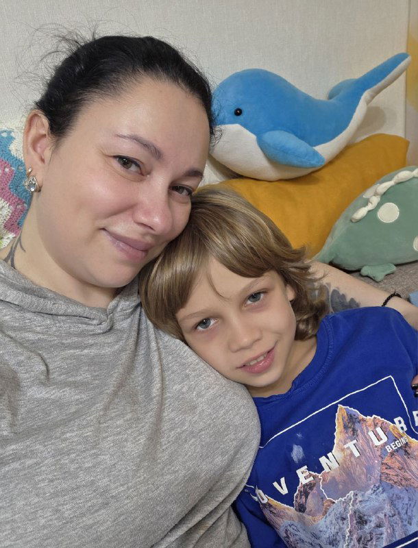 Женщина обнимает ребёнка на диване, оба улыбаются; на фоне мягкая игрушка-дельфин и уютные подушки — семейная сцена дома