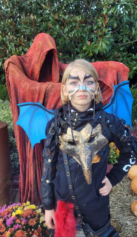 Мальчик в чёрном костюме с синими крыльями и маской позирует на фоне осенней растительности и праздничного оформления парка.